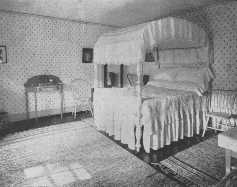 This bed-room is a good example of a simple Colonial bed-room, and the rag rugs are in keeping with it. The repeat design of the wallpaper ties the room into a unified whole.